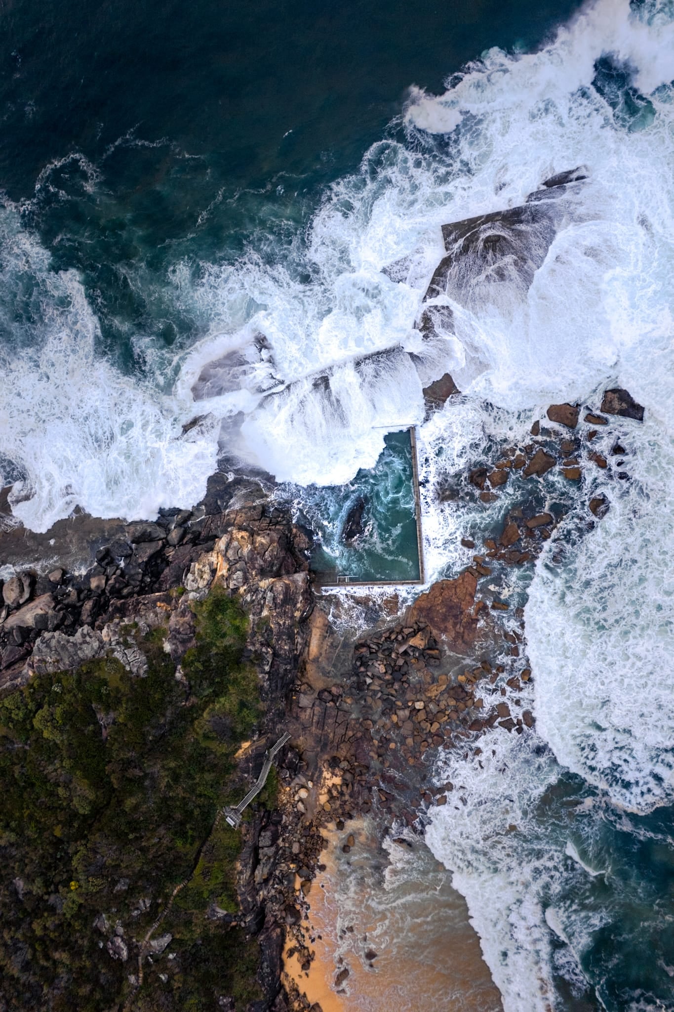 Aerial view of waves crashing against rocky cliffs with a small pool below.
