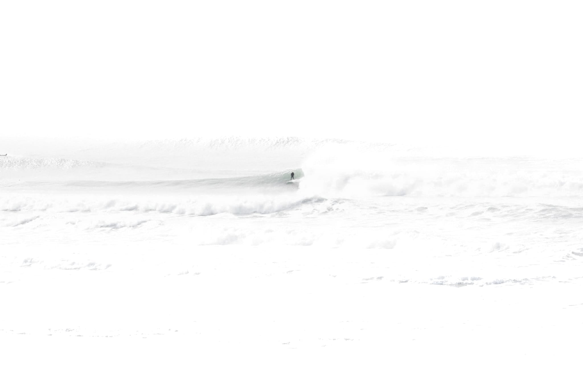 High-key black and white photograph of a lone surfer riding a wave at Ocean Beach, San Francisco, nearly dissolved into white fog and spray.