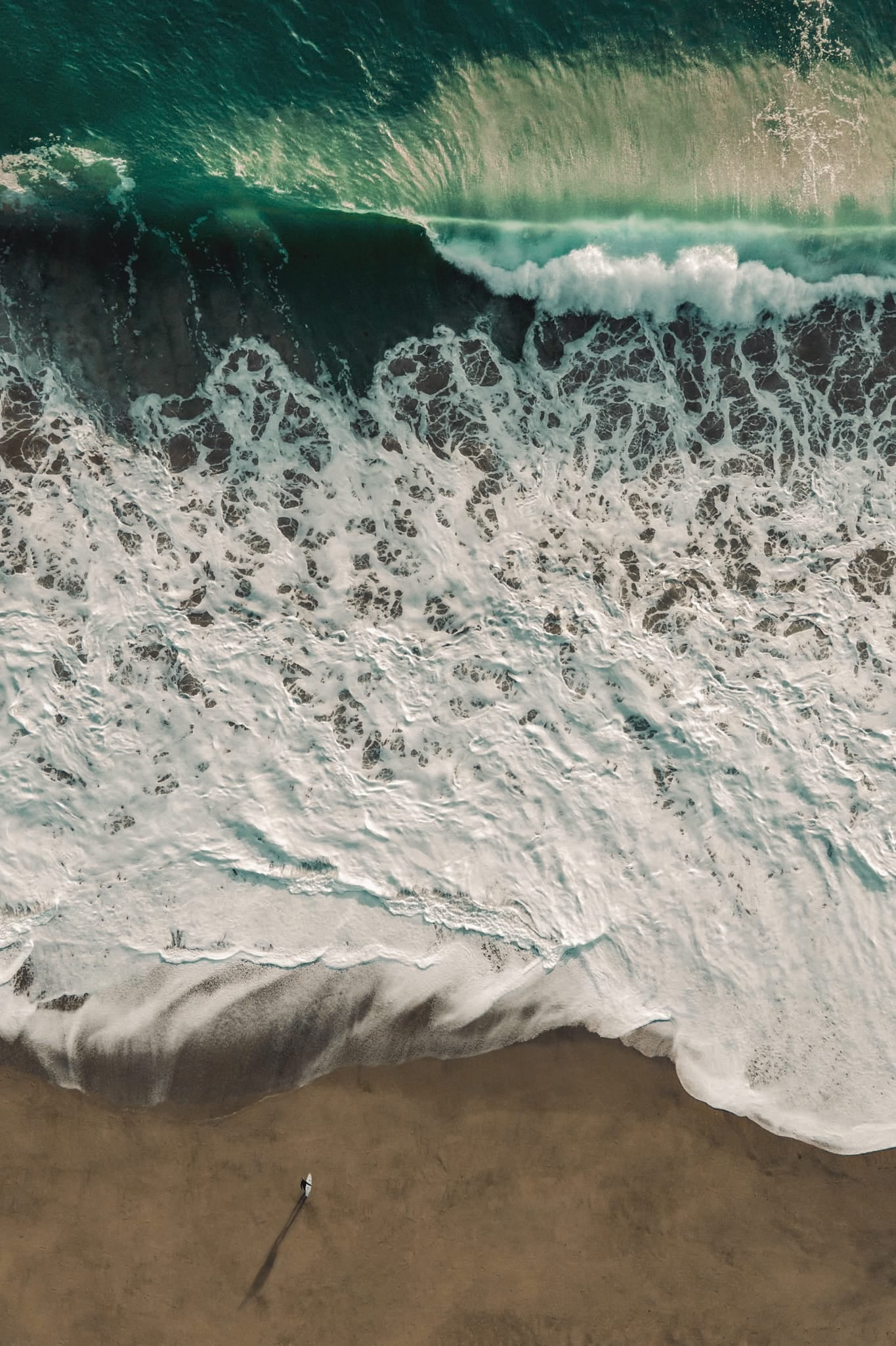 Aerial colour photograph of a lone figure standing on the beach beside lace-like patterns of retreating whitewater, Southern California.