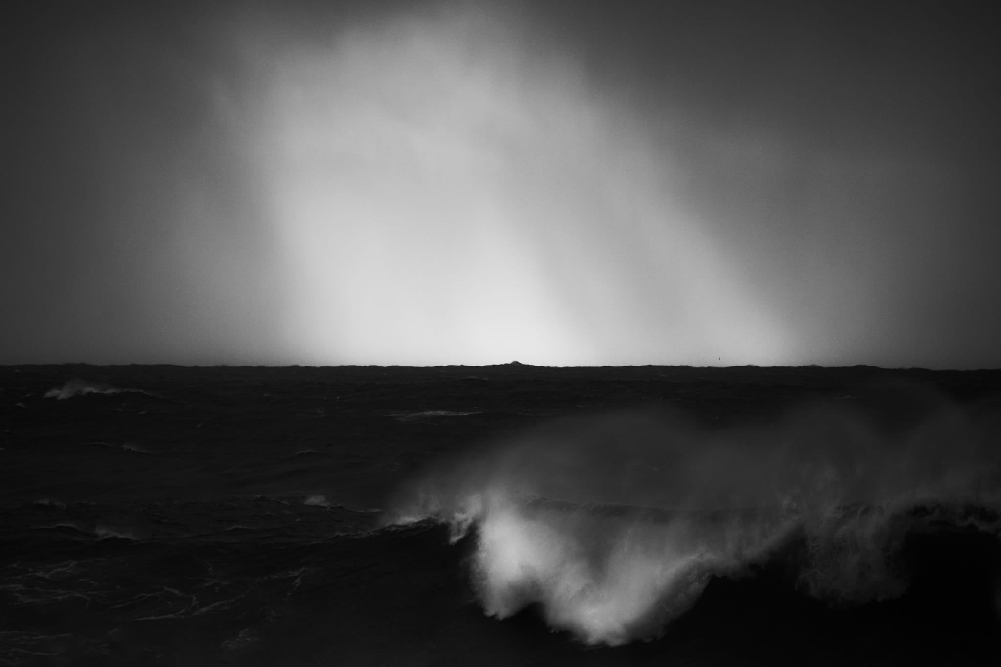 Black and white photograph of dramatic light rays breaking through clouds over a dark wave on the northern Sydney coastline.
