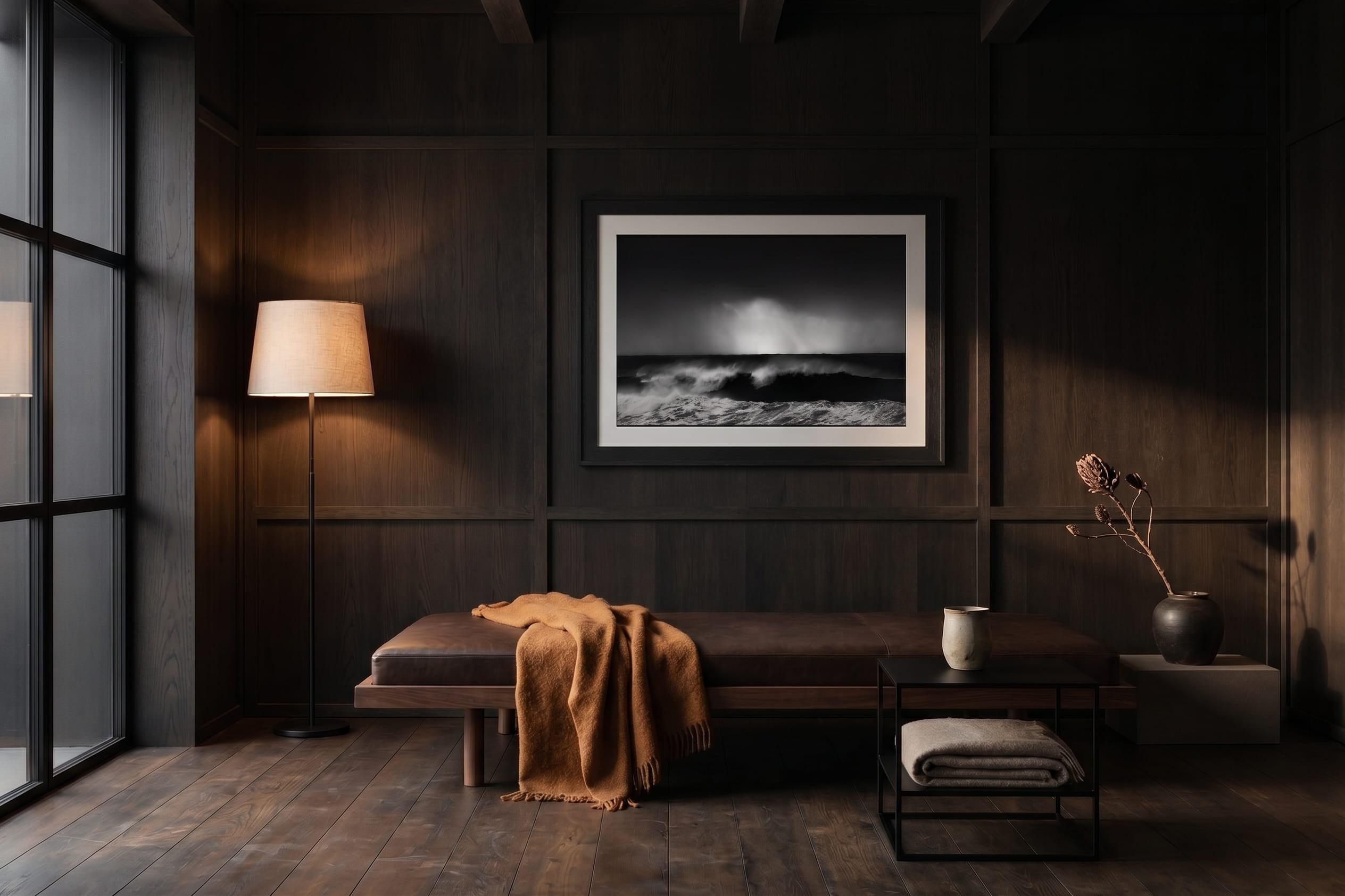 A framed black and white landscape photograph of a stormy ocean wave hangs on a dark wood-paneled wall in a moody, modern living room setting with a bench, floor lamp, and side table.
