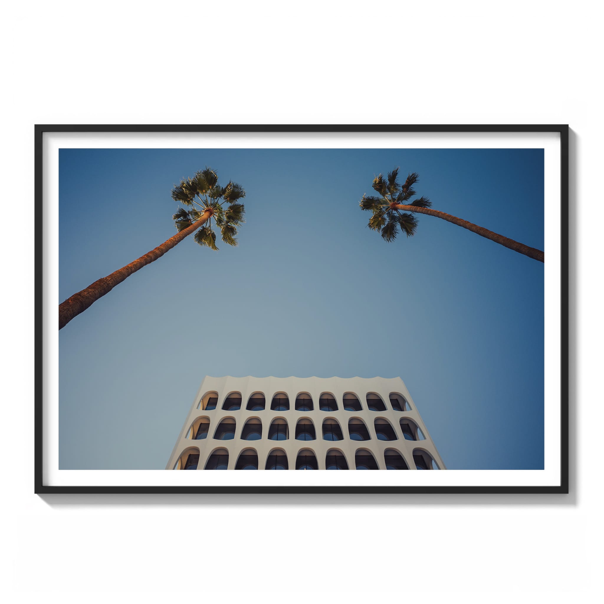 Framed color photograph of two palm trees framing an arched white building facade against a blue sky in Los Angeles.