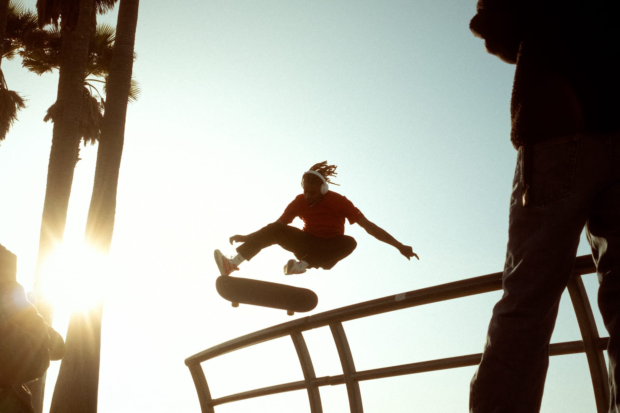 Color photograph of a skateboarder mid-air performing a grab trick, backlit by the sun at Venice Beach with palm trees in the background.