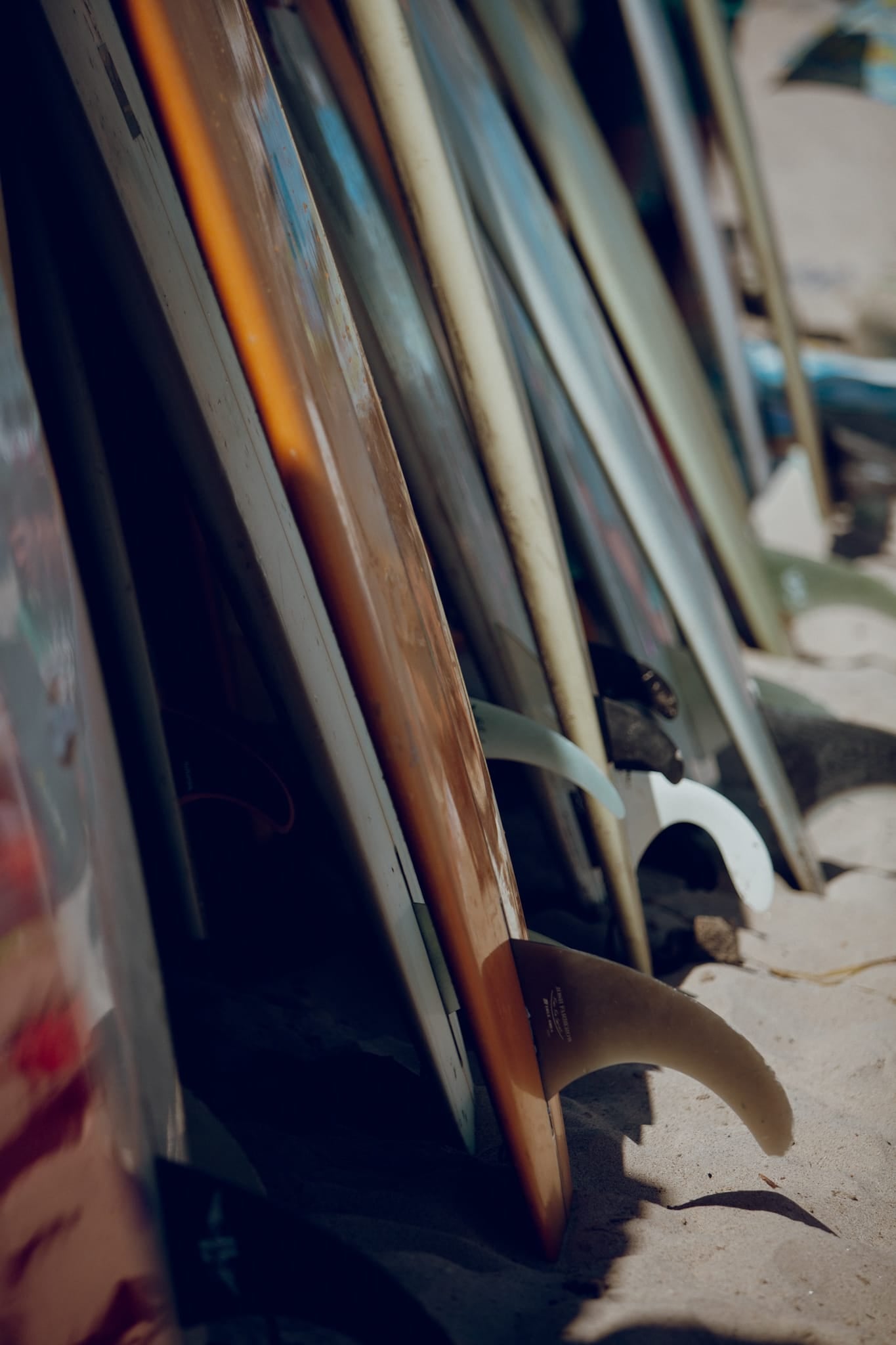 Color photograph of a row of surfboards leaning together on the sand in Malibu, one with a bright orange rail.