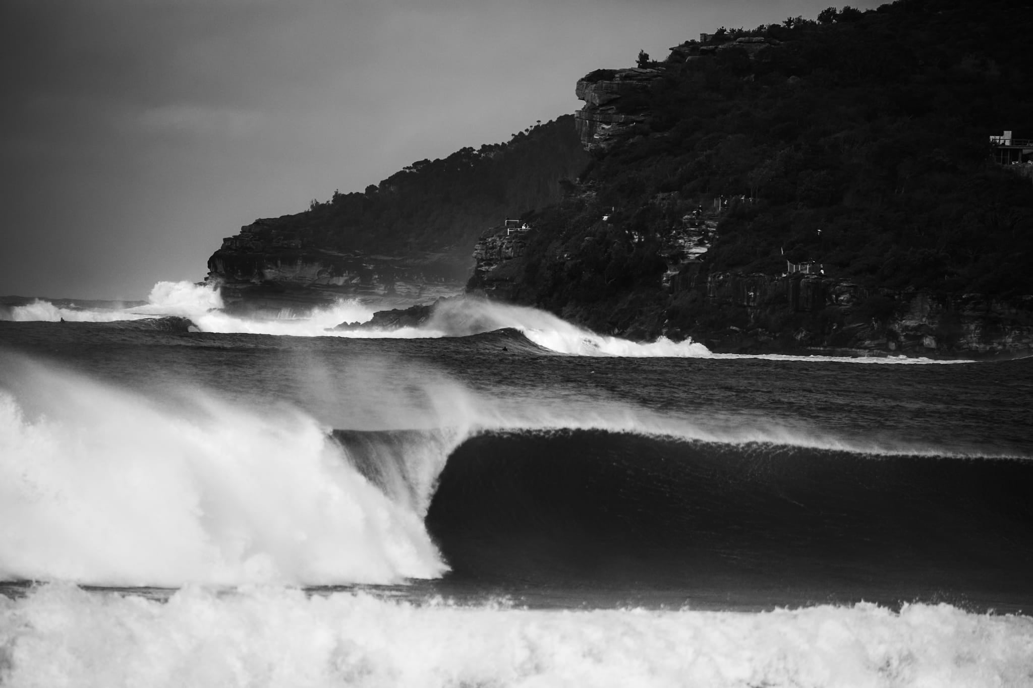 Black and white photograph of a large wave breaking at Manly Beach with North Head visible in the background.