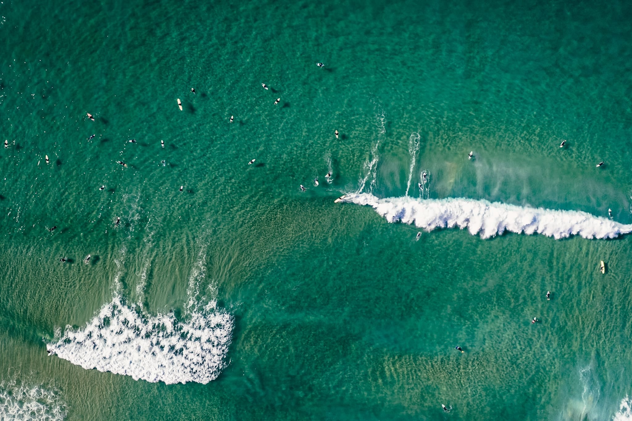 Aerial color photograph of surfers scattered across teal water with branching whitewater patterns at the Northern Beaches, Sydney.