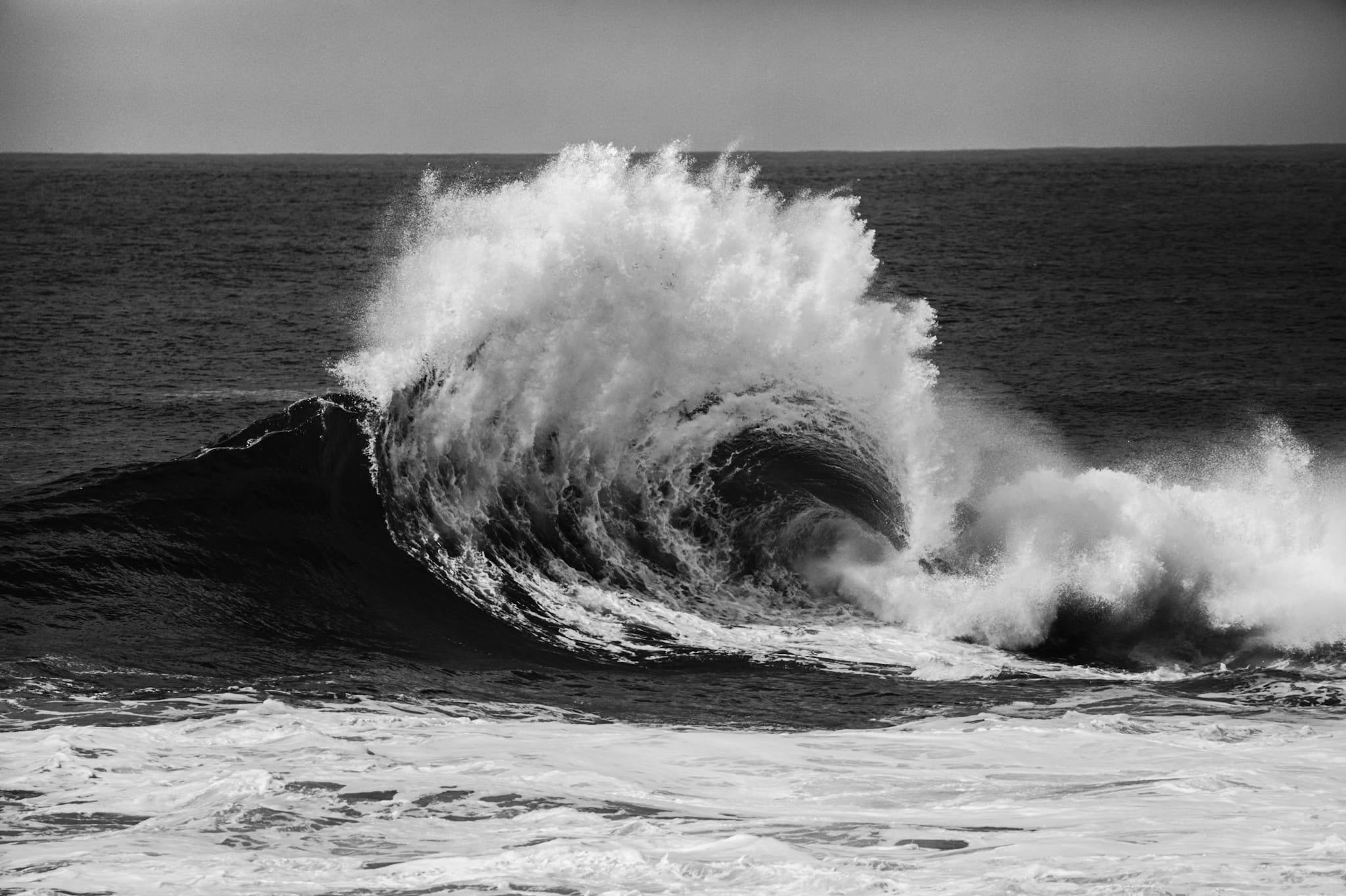 Black and white photograph of a large wave breaking at Cape Solander, spray flaring where backwash off the rock shelf collides with the incoming swell