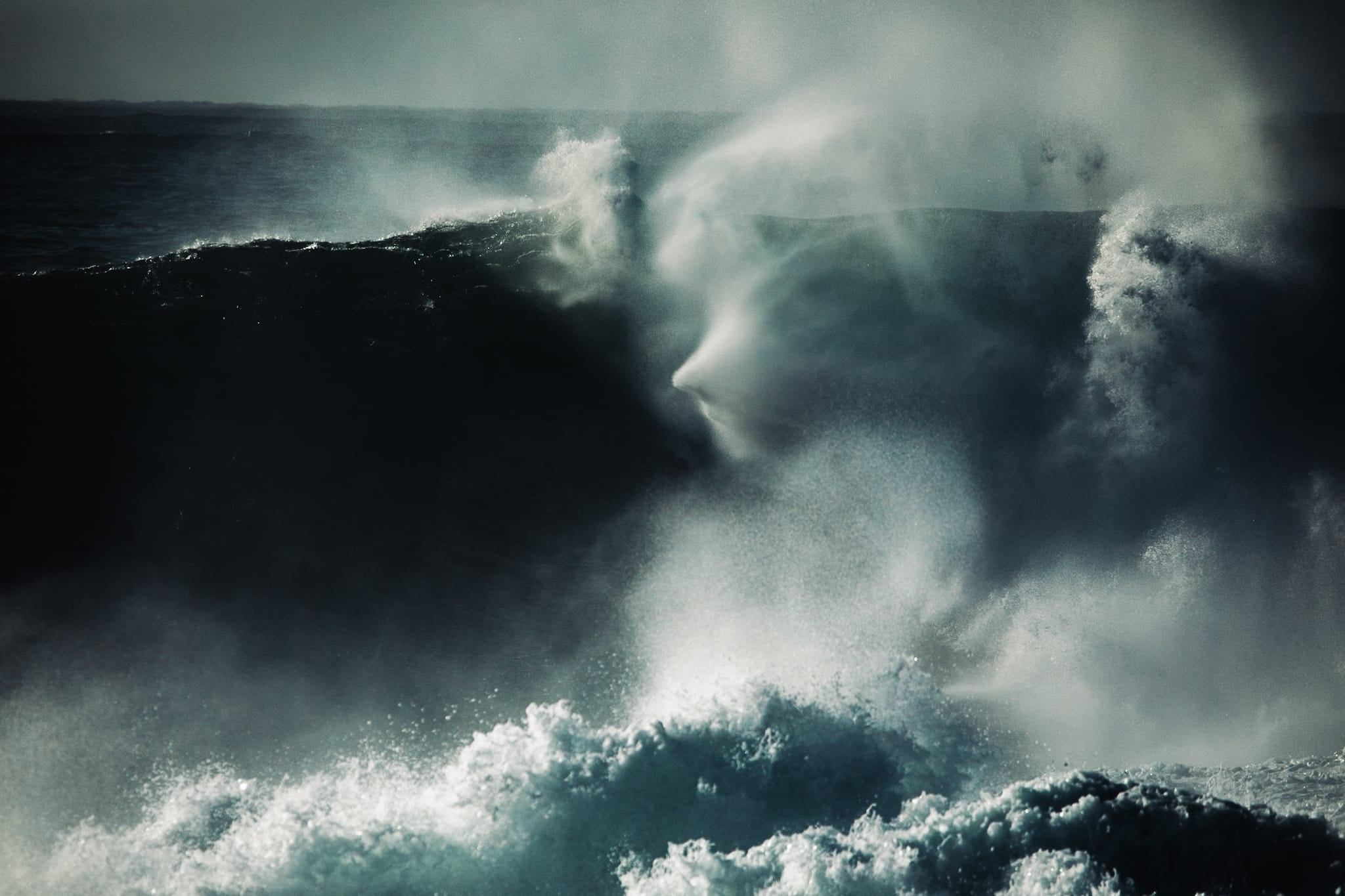 Black and white photograph of a heavy slab wave breaking near Sydney, with mist forming an eerie face-like shape above the barrel.