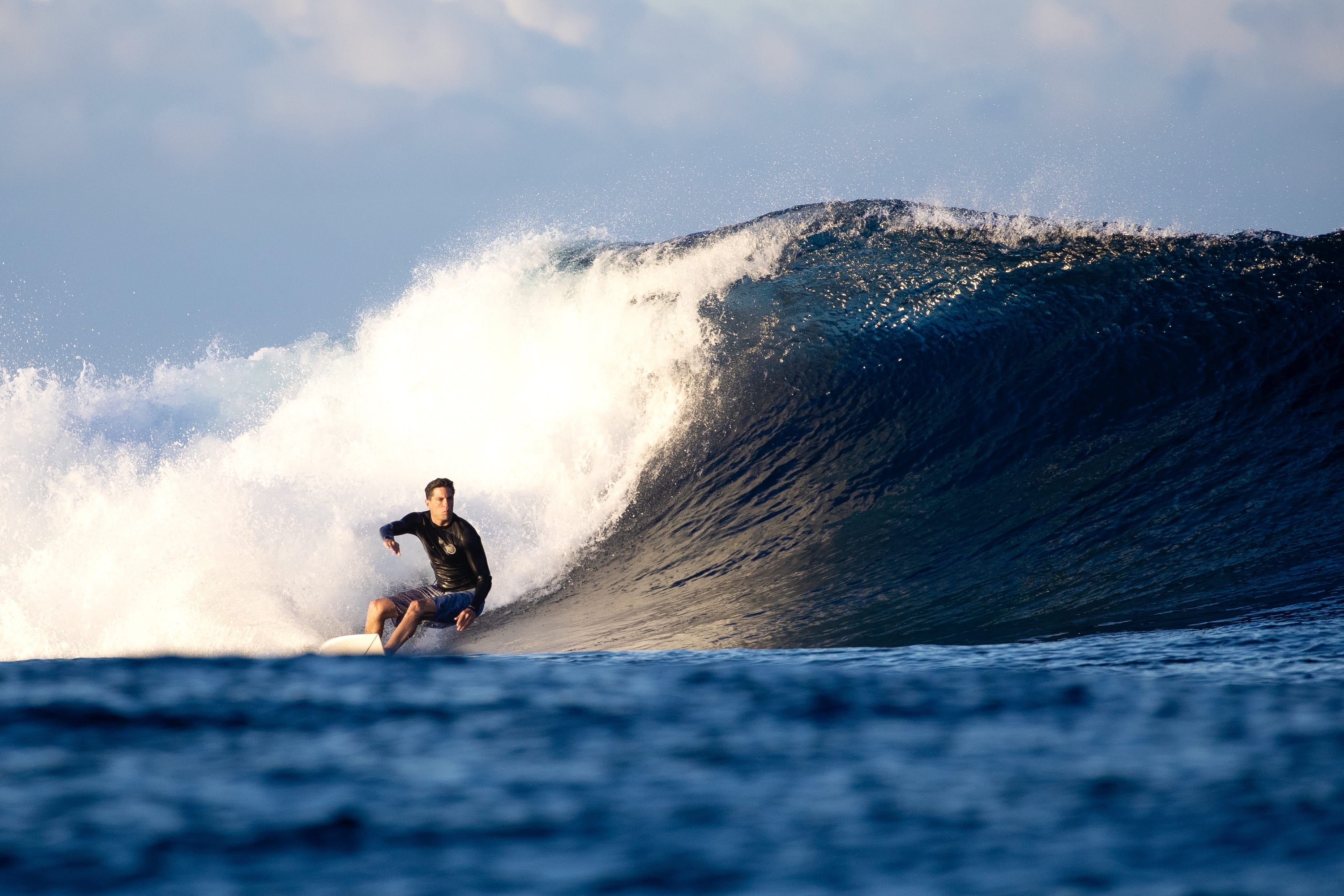 scott gudahl cloudbreak fiji, a large curling ocean wave under a blue sky, with white spray and deep blue water visible.