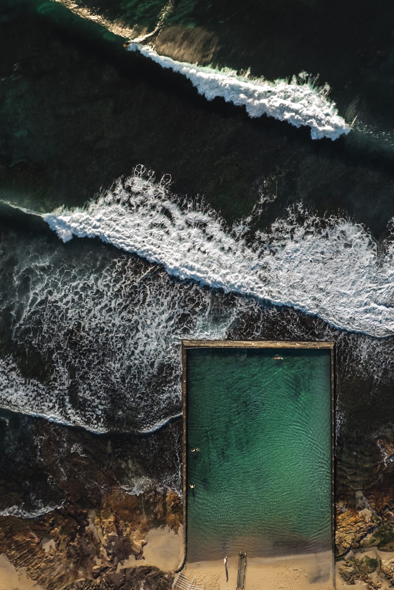 Aerial color photograph of a rectangular ocean rock pool beside crashing waves at Cronulla at sunrise.