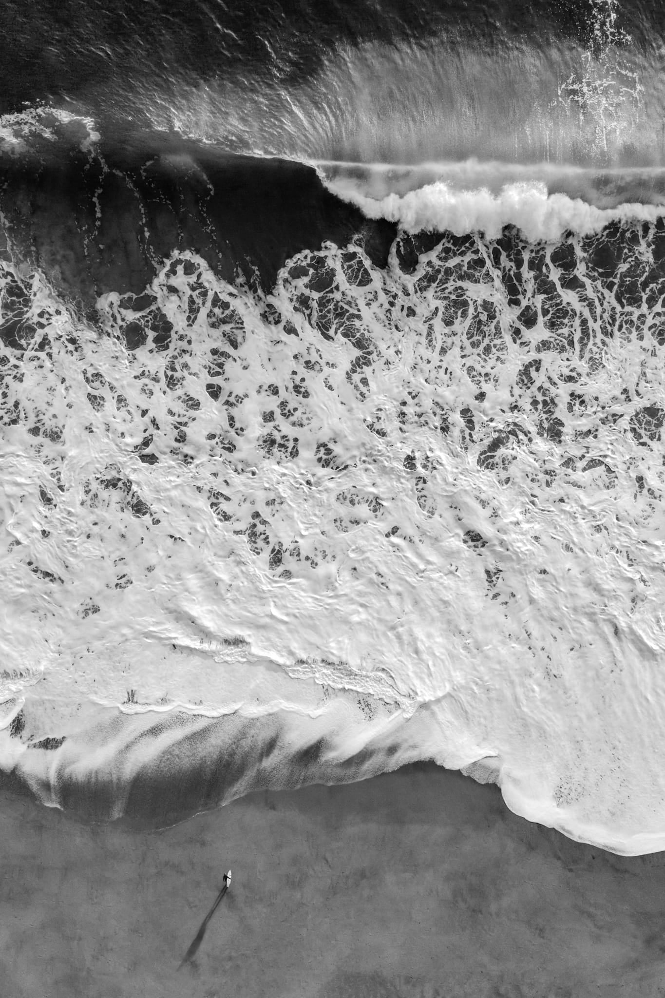 Aerial black and white photograph of a lone figure standing on the beach beside lace-like patterns of retreating whitewater, Southern California.