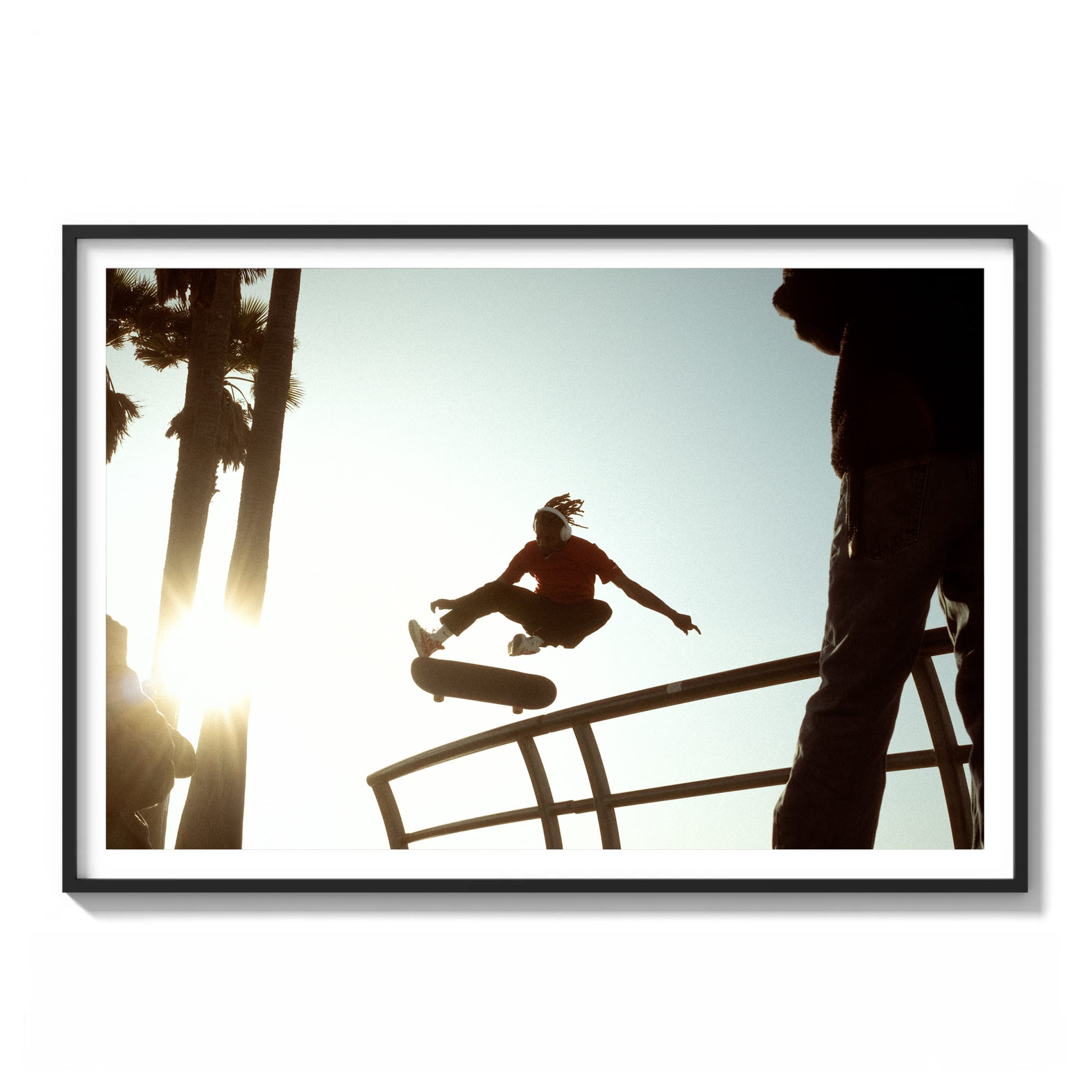 Framed color photograph of a skateboarder mid-air performing a grab trick, backlit by the sun at Venice Beach with palm trees in the background.