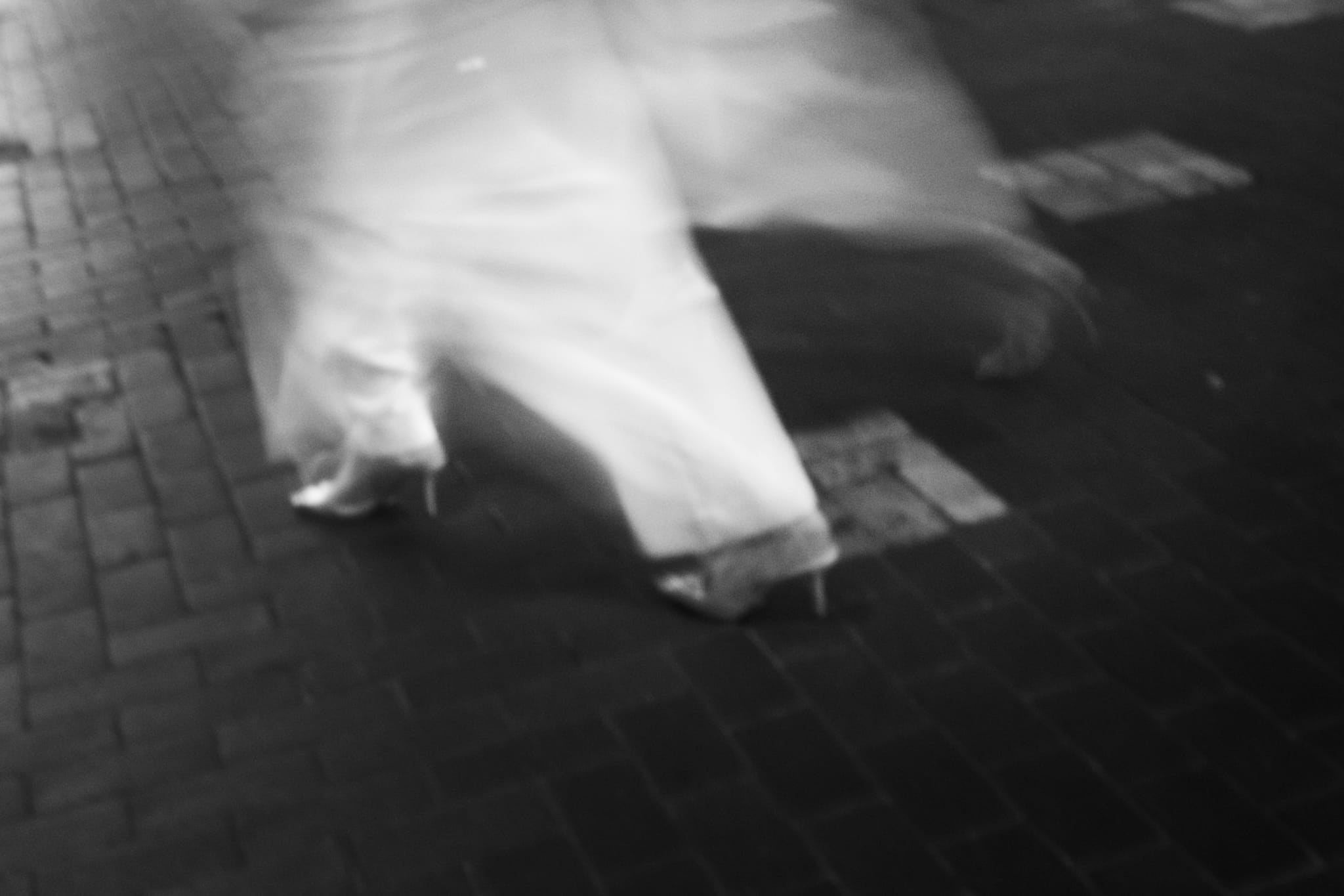 Black and white long-exposure photograph of a blurred figure in a white dress walking on cobblestone in the Sydney CBD at night.