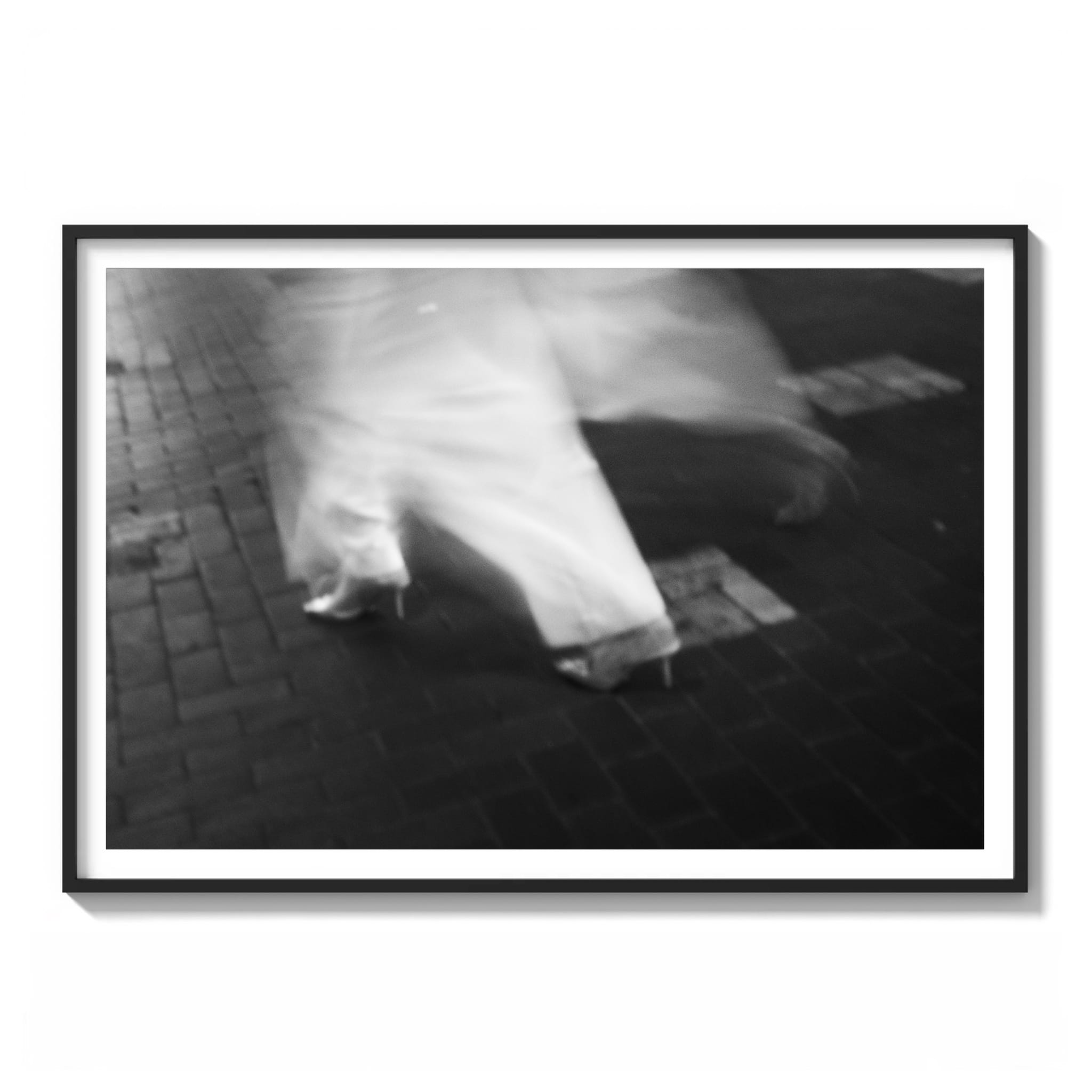 Framed black and white long-exposure photograph of a blurred figure in a white dress walking on cobblestone in the Sydney CBD at night.