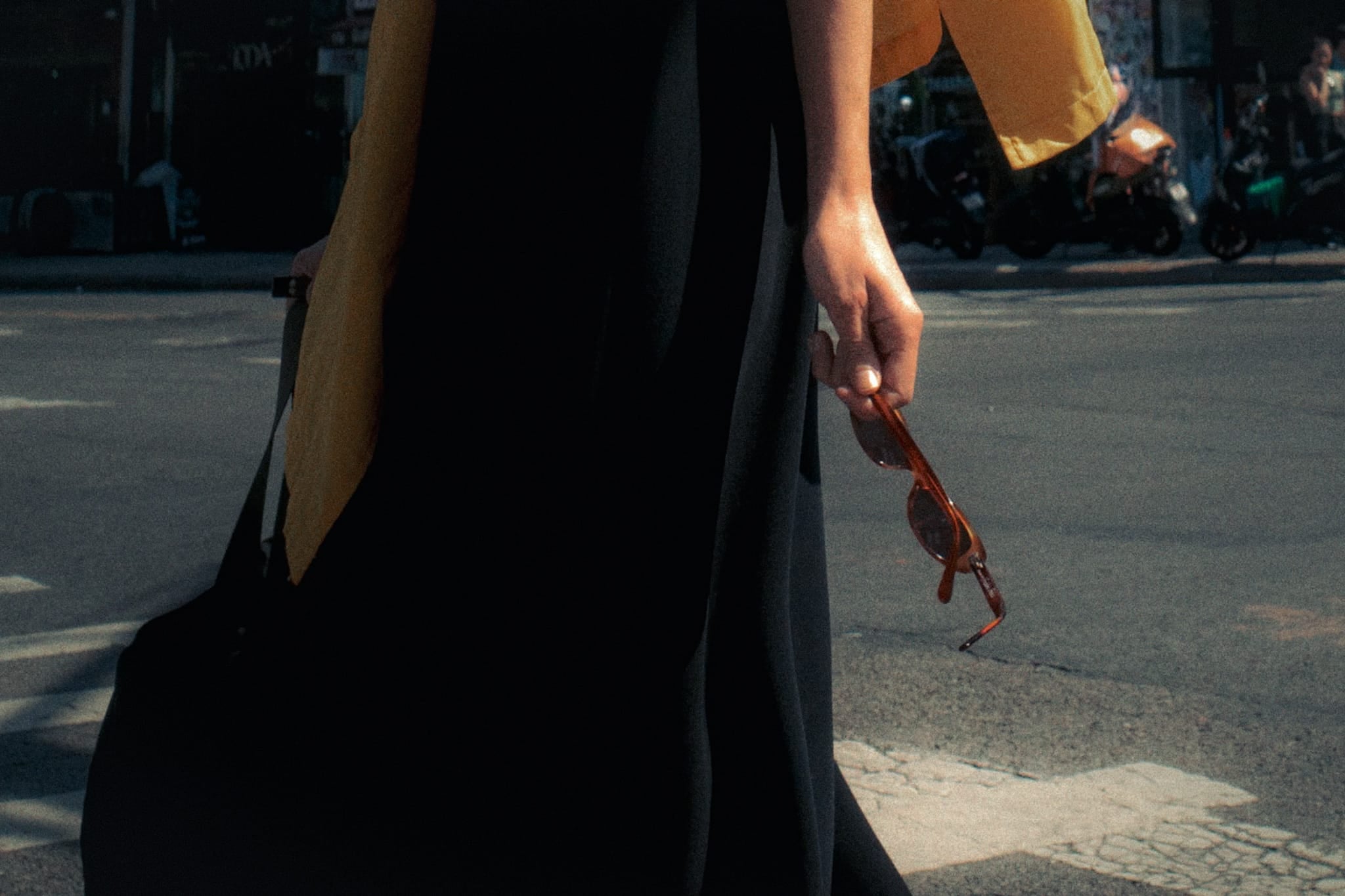 Color street photograph of a person in a black dress and mustard scarf holding sunglasses while crossing a street in Brooklyn, New York.