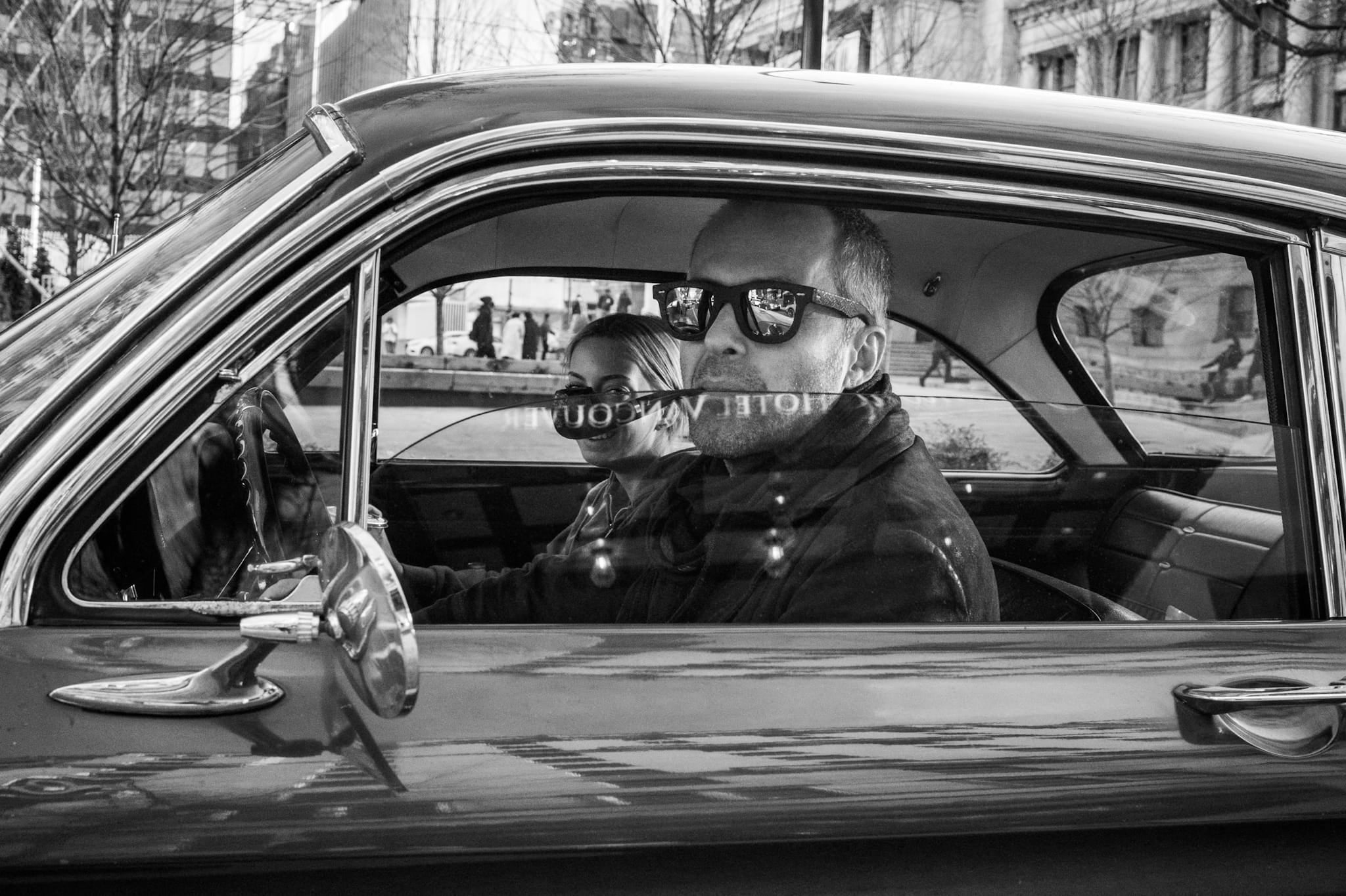 Black and white photograph of a couple sitting in a vintage car with "Hotel Vancouver" reflected across the windshield.