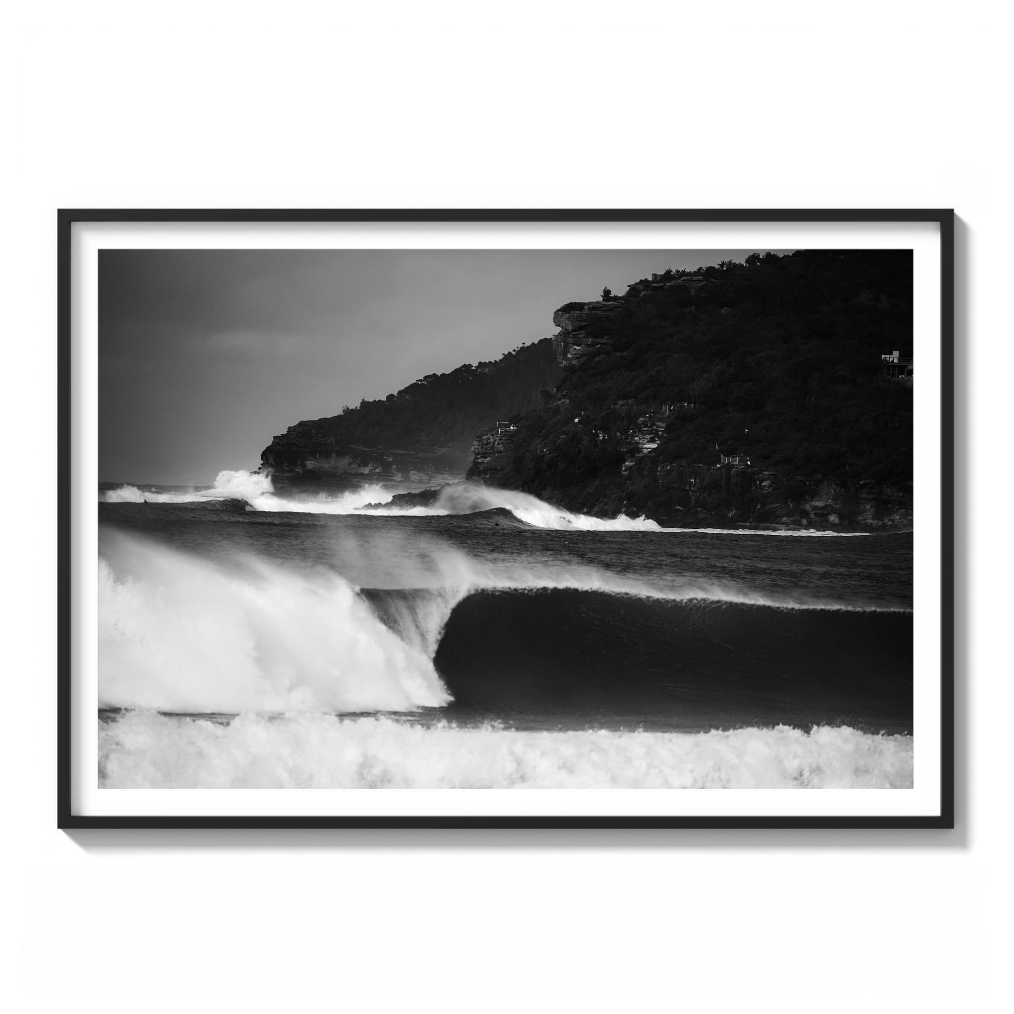 Framed black and white photograph of a large wave breaking at Manly Beach with North Head visible in the background.