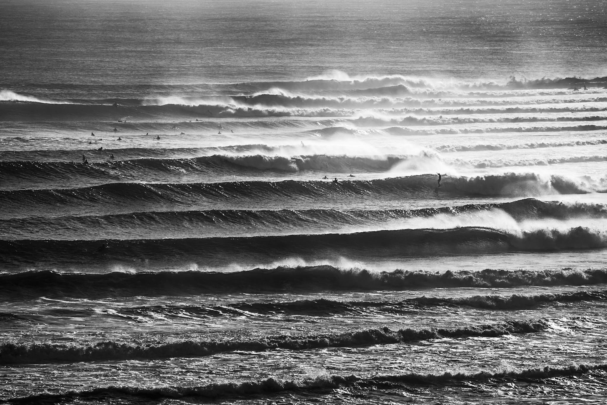 Black and white photograph of multiple parallel wave lines breaking along the Gold Coast with surfers scattered across the lineup.